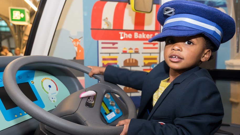Young child in driver's cap holding a steering wheel at London Transport Museum