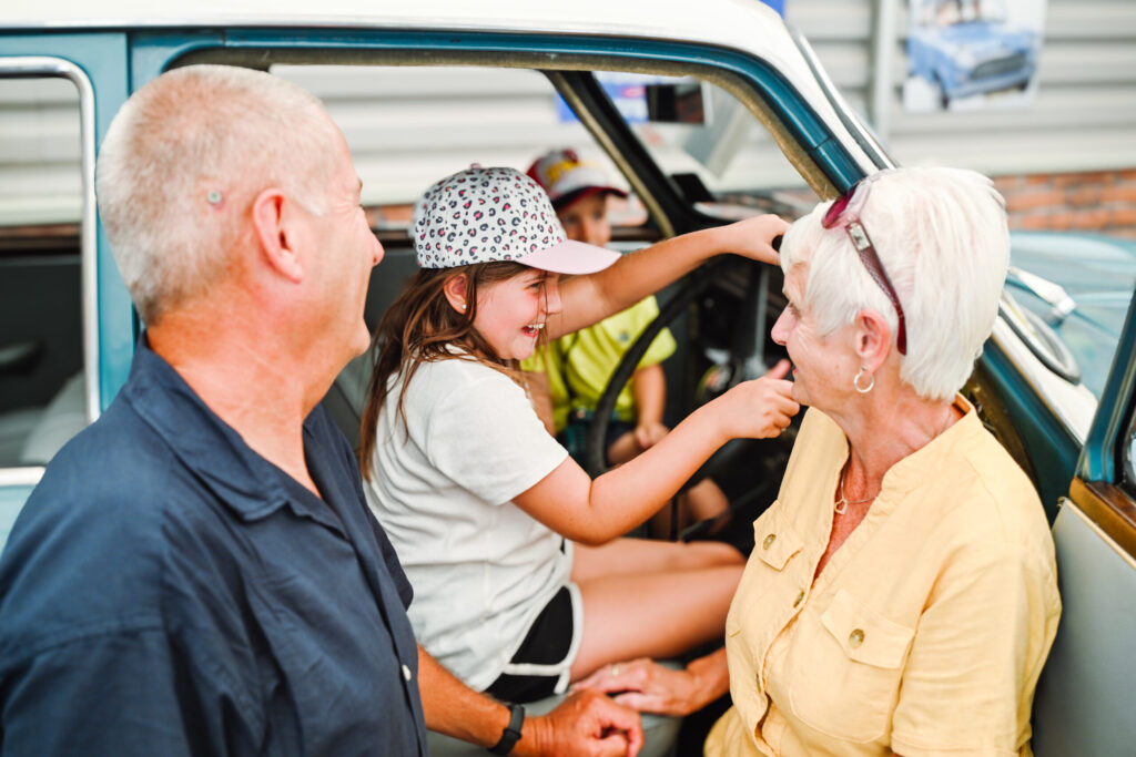 girl with white and black baseball cap sits in a seat of an old car with a grandma and grandad standing outside