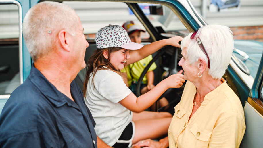 girl with white and black baseball cap sits in a seat of an old car with a grandma and grandad standing outside