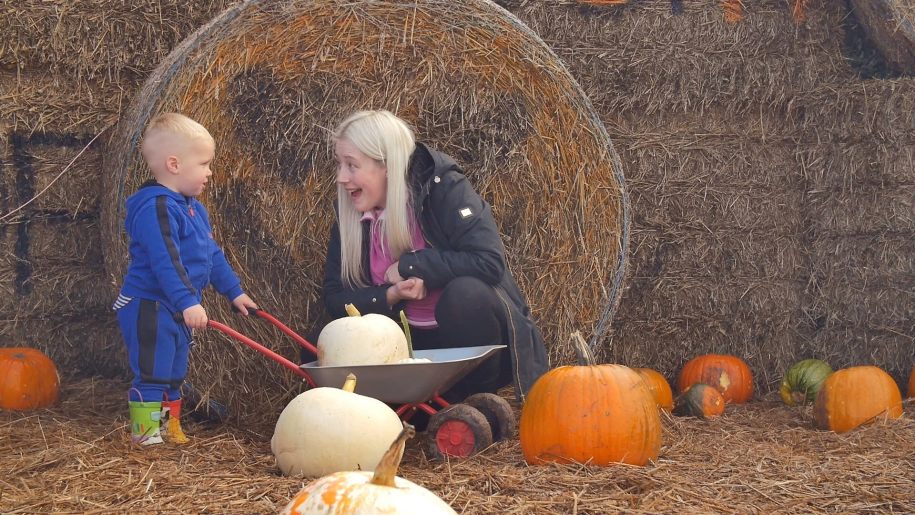 adult smiling at young child pushing a wheelbarrow containing a pumpkin