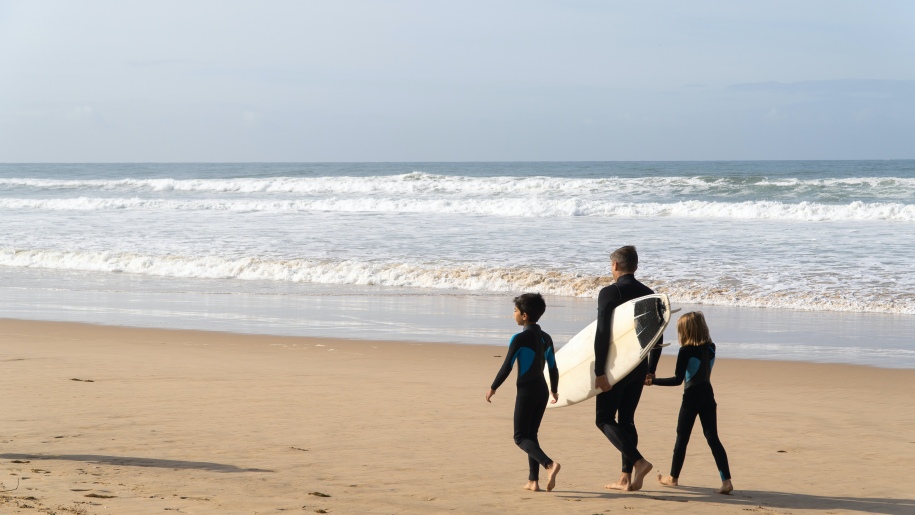 Adult and two children with a surfboard on a beach.