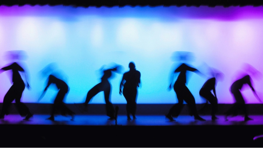 Silhouettes of dancers on a stage illuminated in blue and purple light.