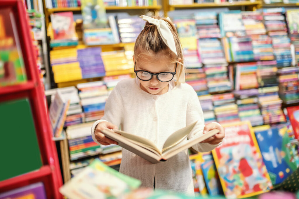Adorable curious caucasian little girl with eyeglasses standing in bookstore and reading interesting book. All around are books on shelves.