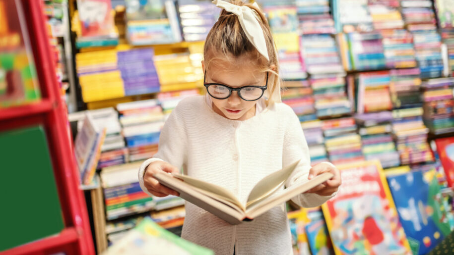 Adorable curious caucasian little girl with eyeglasses standing in bookstore and reading interesting book. All around are books on shelves.