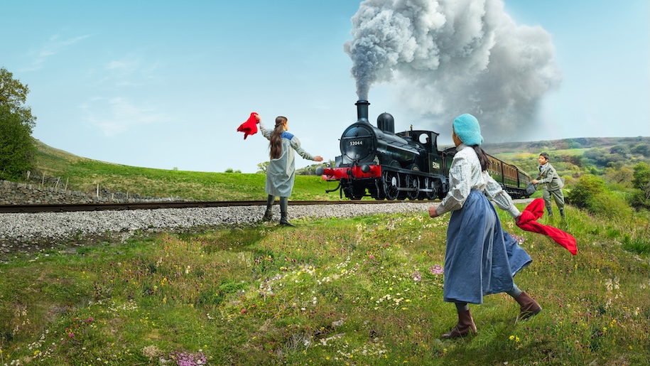 girl waves a red flag as a steam train goes through a field