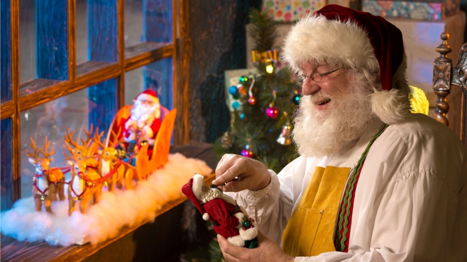 Santa Claus in his workshop looking at a toy Santa.