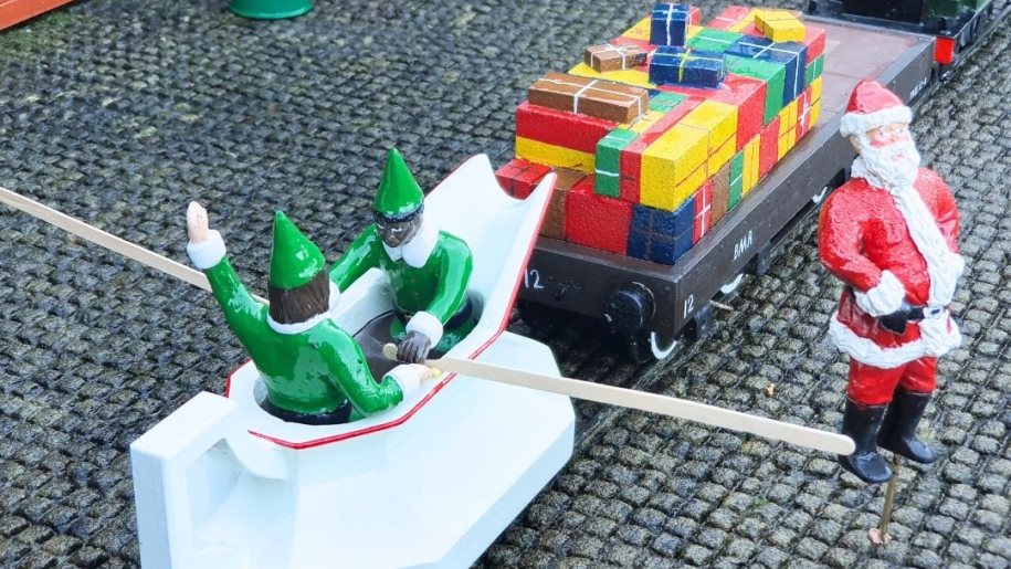 A little model Santa standing beside a wagon loaded with Christmas presents.