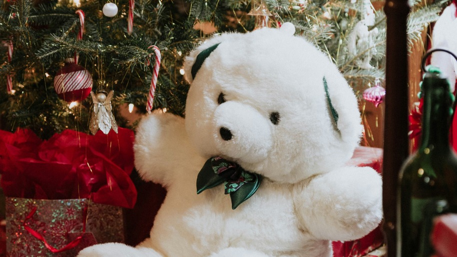 A white teddy bear with a green bow by a Christmas tree.
