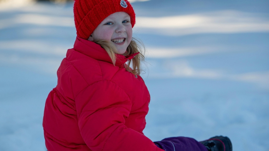 Happy child in red hat and jacket sitting in snow.