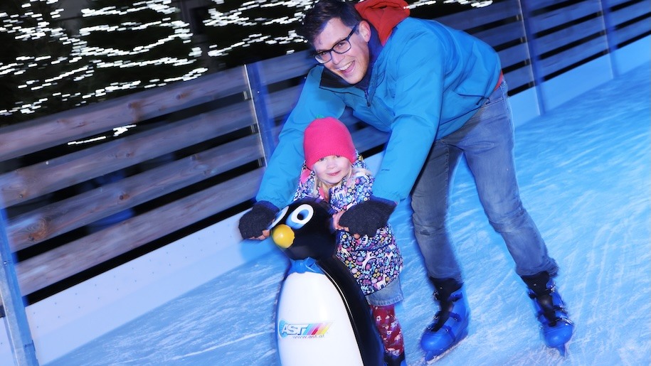Father, young daughter and penguin skate aid on an outdoor ice rink.