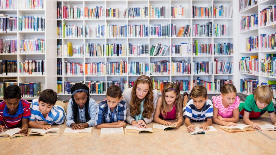 line of children laying on their fronts reading with bookcases behind them