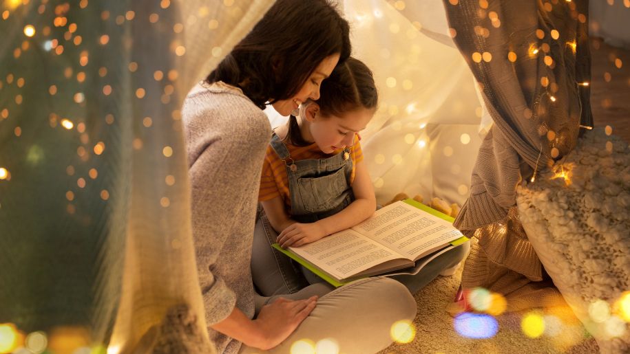 girl reads in fairy light covered tent with mum