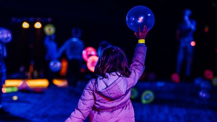 Child in a purple jacket holding up a ball at an illuminated funfair.