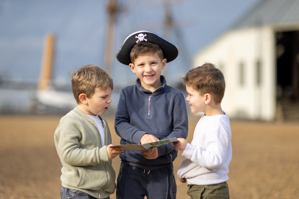 three boys, one in a pirate hat standing on a beach looking at a map