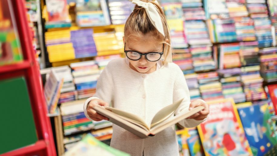 Adorable curious caucasian little girl with eyeglasses standing in bookstore and reading interesting book. All around are books on shelves.