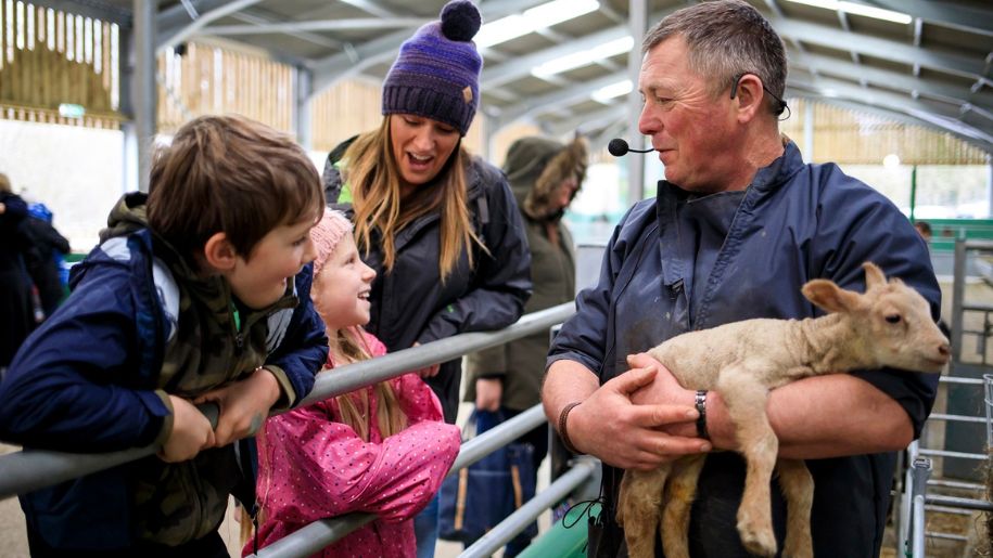farmer holds newborn lamb while family smile and look at it over gate