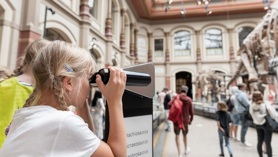 girl looks through binoculars at a museum behind her