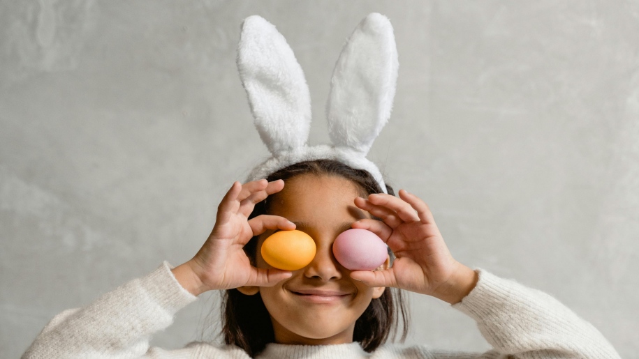 Girl wearing white bunny ears holding two Easter eggs over her eyes.
