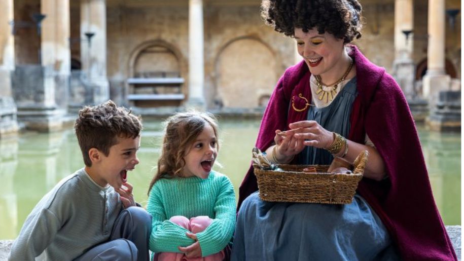 lady dressed in purple robe by roman baths talks to two children 