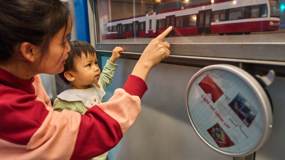a mother in a pink and red striped jumper holds a child and points at transport exhibit