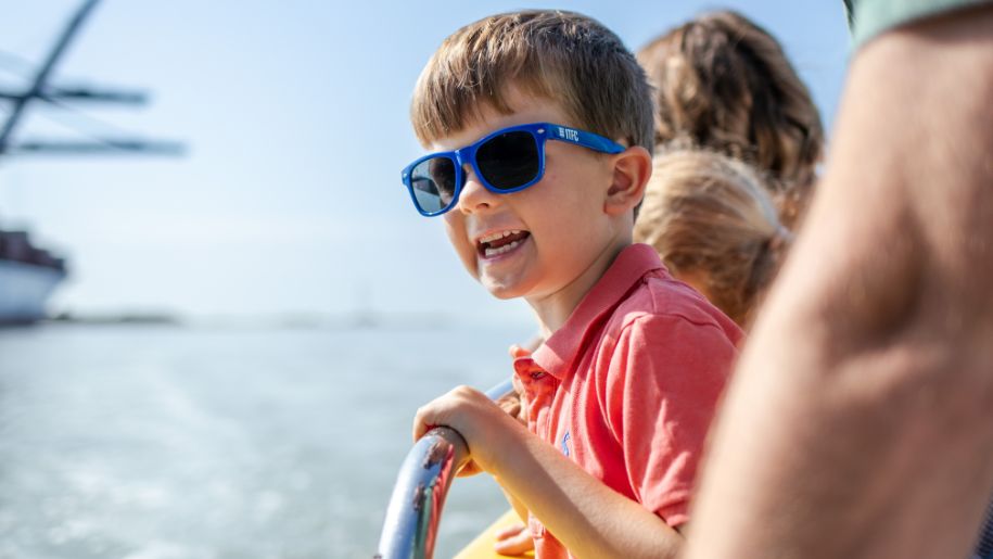 boy in red tshirt and blue sunglasses smiles while looking over edge of yellow boat onto sea