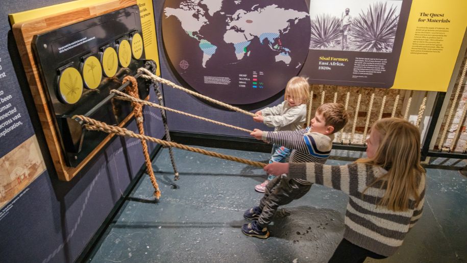 three children pull on ropes attached to yellow and black exhibits onn wall of the world and plants