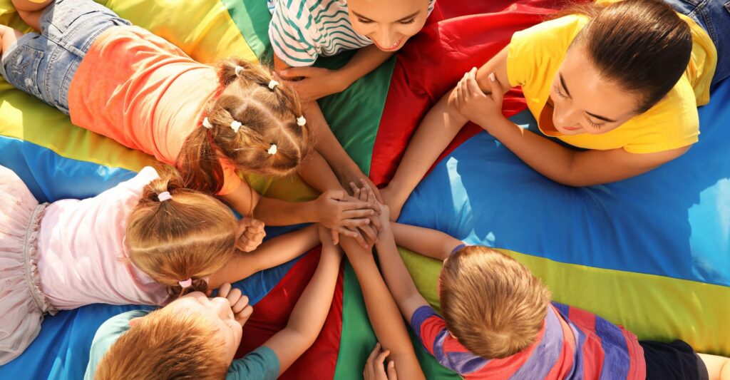Group of children with teachers holding hands together on rainbow playground parachute, top view. Summer camp activity