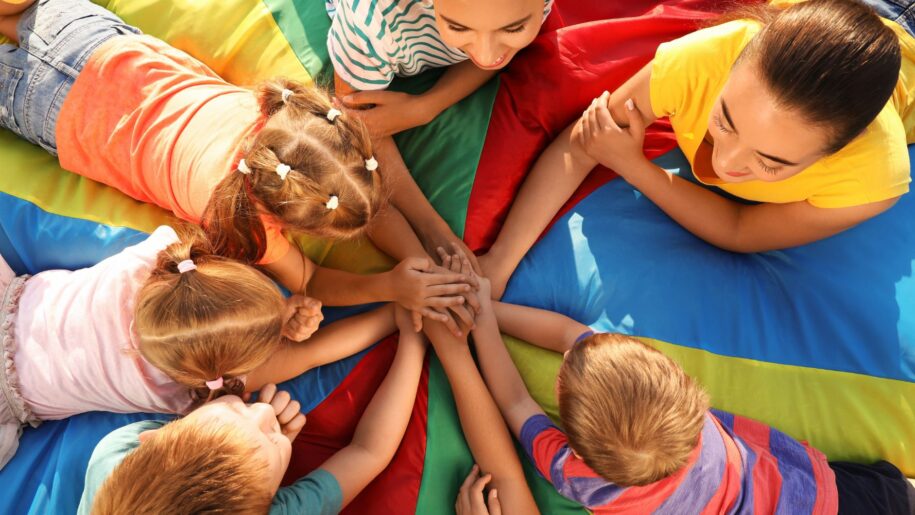 Group of children with teachers holding hands together on rainbow playground parachute, top view. Summer camp activity