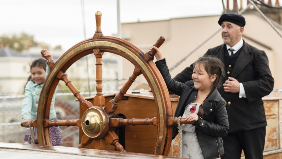 A costumed character watching two children turn a ship's wheel.