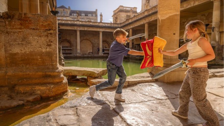two children pretend to battle with swords infront of a roman pillar and bath