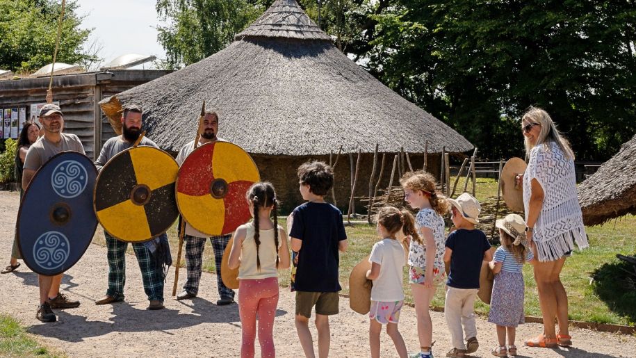 Celtic warriors and a group of children standing by a thatched hut.