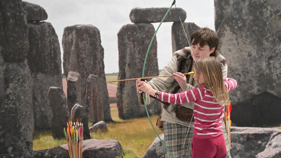 Adult showing a child how to fire a bow and arrow against a backdrop of standing stones.