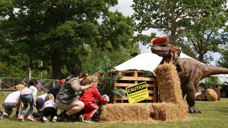 Children watching a large model dinosaur from behind strawbales.