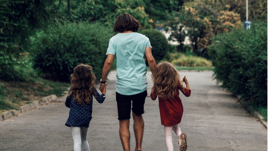 Adult and two young girls holding hands as they run down a path between shrubs.