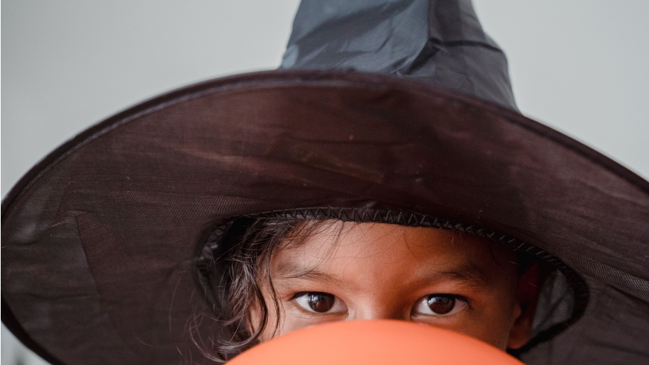Child in a black witch's hat peeping over an orange balloon.