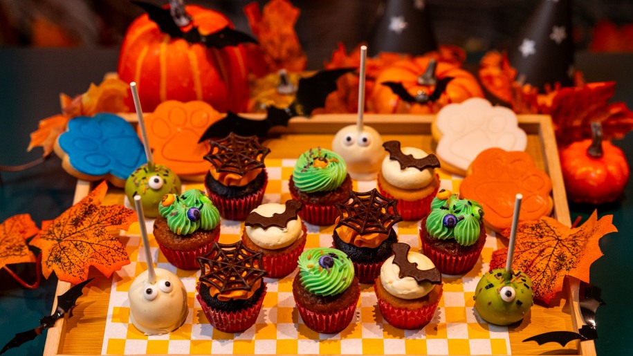 A tray of Halloween-themed cup cakes with autumn leaves, bats and pumpkins in the background.